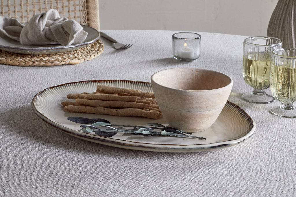 The nkuku Malia Large Oval Platter in Cream displays breadsticks, a wooden bowl, and greenery on a textured tablecloth. Around it are two sparkling drinks in glasses, a small candle, and a plate with a napkin.