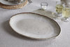 The nkuku Malia Large Oval Platter in cream, featuring an organic shape and textured rim, sits on a beige tablecloth beside two sparkling drinks, a small candle, and a plate with a napkin.