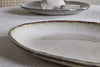 A close-up of the nkuku Malia Large Oval Platter in cream, featuring a textured, ridged rim and organic shape on a light tablecloth, with another platter and a folded napkin blurred in the background.