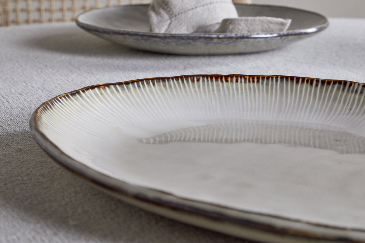 A close-up of the nkuku Malia Large Oval Platter in cream, featuring a textured, ridged rim and organic shape on a light tablecloth, with another platter and a folded napkin blurred in the background.