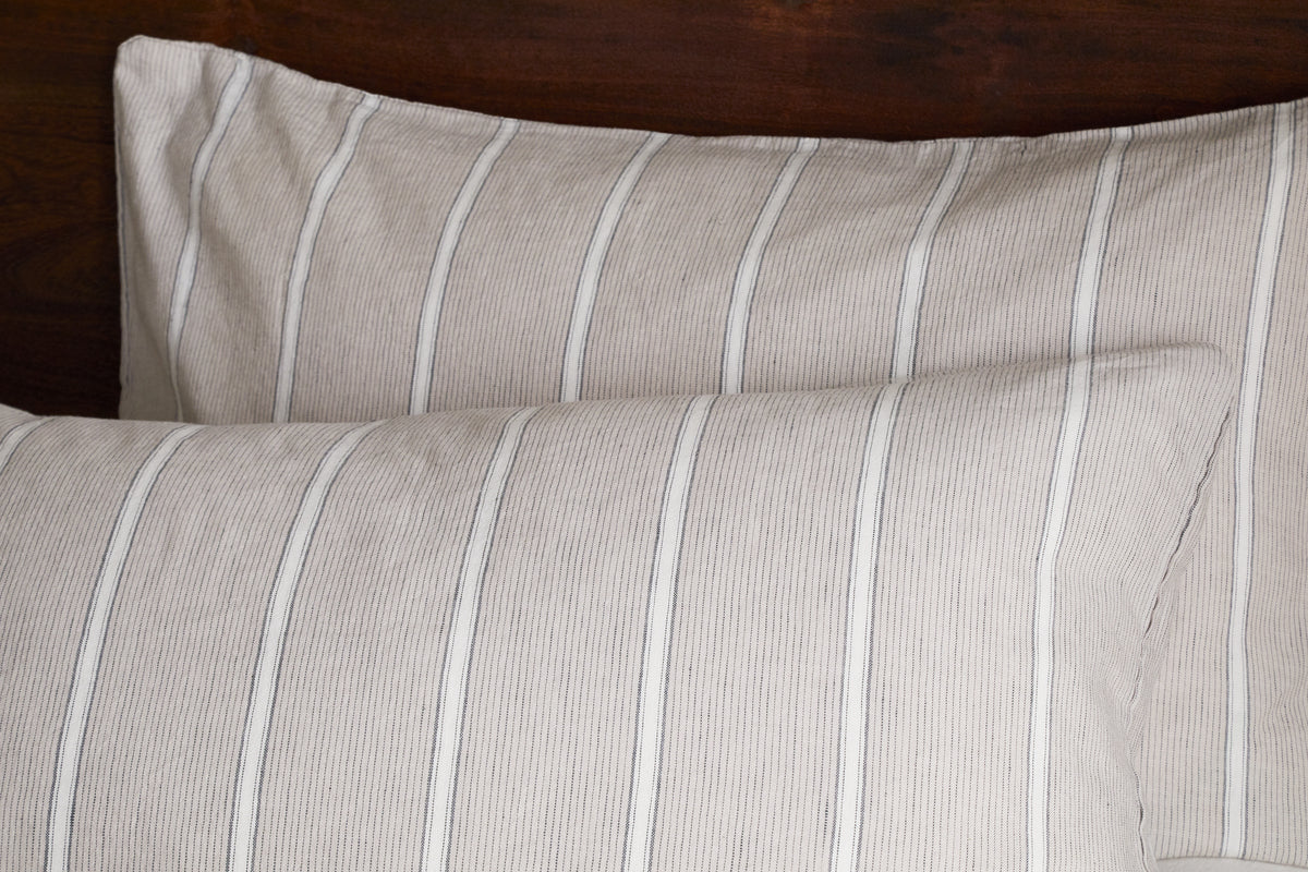 Two pillows with Mora Hemp & Cotton Housewife Pillowcases in Stone Grey by nkuku rest against a dark wooden headboard, featuring soft, comfortable fabric with subtle thin stripes.