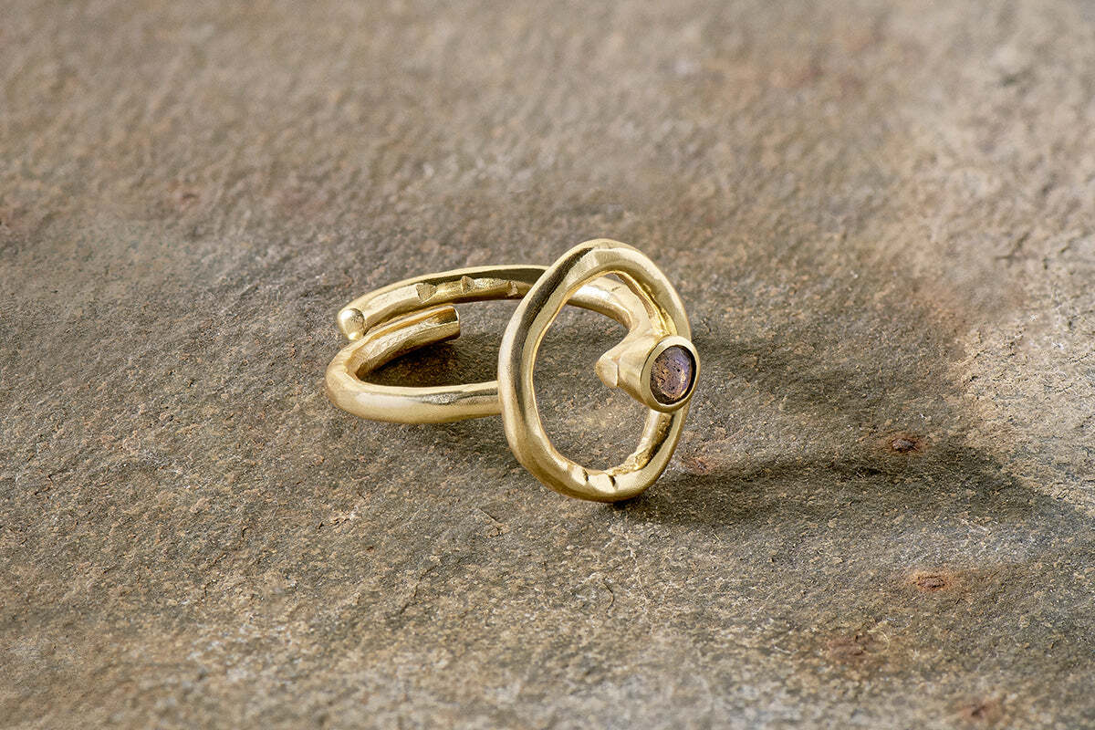 The Mari Labradorite Ring - Gold from nkuku features a hypoallergenic, gold-plated adjustable open band with an irregular circular design and small labradorite gemstone, displayed on a textured stone surface.