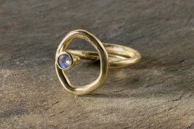 The nkuku Mari Labradorite Ring - Gold features a gold plated, organic open circle design with a bezel-set iridescent Labradorite stone, a matte hypoallergenic finish, and is shown on textured stone.
