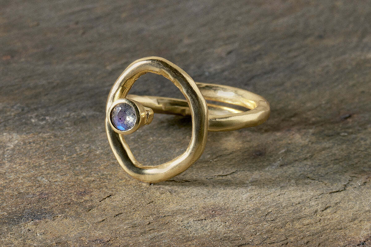 The nkuku Mari Labradorite Ring - Gold features a gold plated, organic open circle design with a bezel-set iridescent Labradorite stone, a matte hypoallergenic finish, and is shown on textured stone.