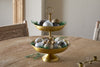 The Mahika Stand - Antique Brass by nkuku is a two-tiered bowl that makes a stunning centrepiece, ideal for festive tablescapes when adorned with ornaments and green leaves.