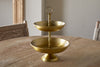 The Mahika Stand - Antique Brass by nkuku, a two-tiered gold metal serving tray ideal as a centerpiece or cake stand, is displayed on a round wooden table with woven chairs in the background.