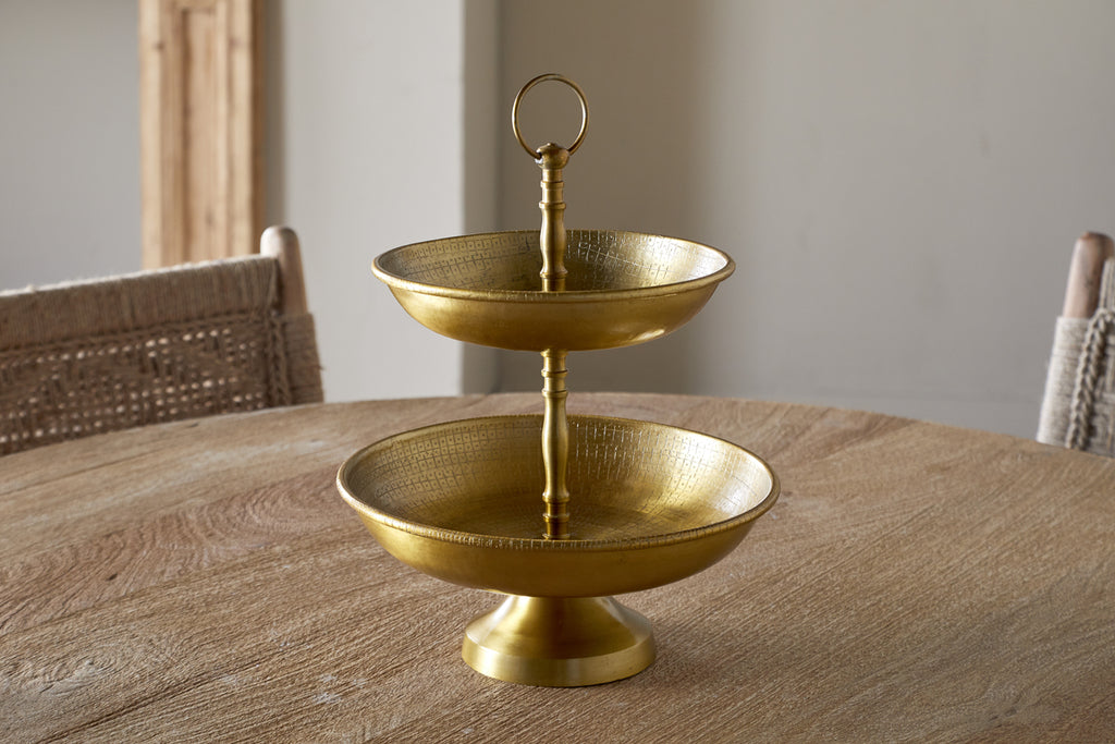The Mahika Stand - Antique Brass by nkuku, a two-tiered gold metal serving tray ideal as a centerpiece or cake stand, is displayed on a round wooden table with woven chairs in the background.