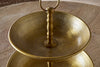 A close-up of the Mahika Stand - Antique Brass by nkuku, a two-tiered brass serving tray with ornate patterns and a central handle, ideal as a festive table centrepiece. Placed on a wooden surface.