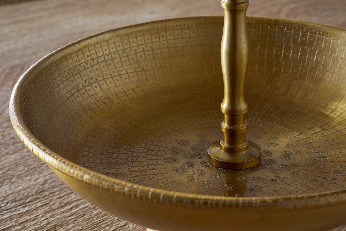 A close-up of the Mahika Stand - Antique Brass by nkuku, featuring engraved patterns and a central pedestal, perfect as a festive table centrepiece, displayed on a wooden surface.