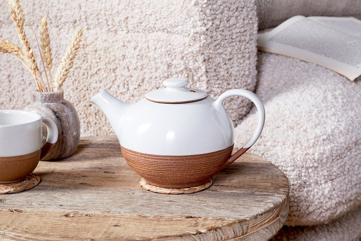 The Mali Ribbed Teapot - White by nkuku, with its brown textured base, sits on a wooden table next to a matching teacup, a vase of wheat stalks, and an open book resting on a soft cream chair in the background.