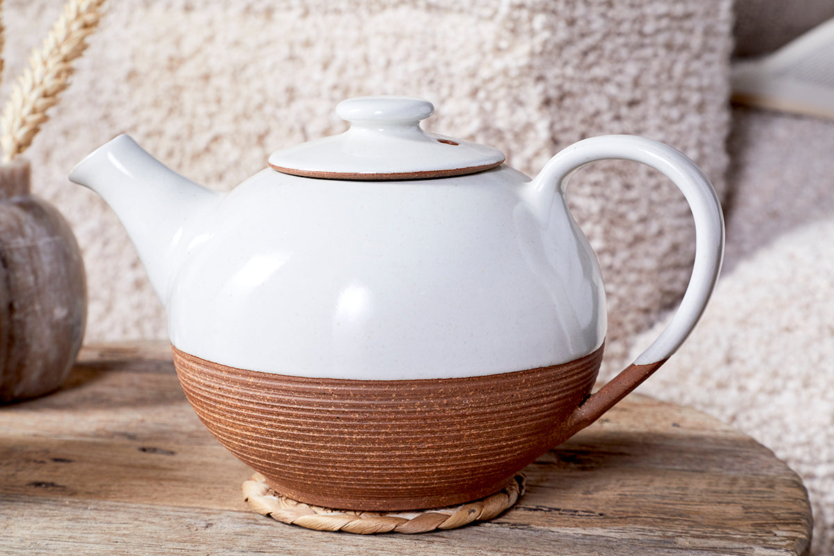 The Mali Ribbed Teapot - White by nkuku, featuring a white glazed top and textured brown base, rests on a woven coaster on a rustic wooden table with soft beige fabric and a vase of dried grass in the background.