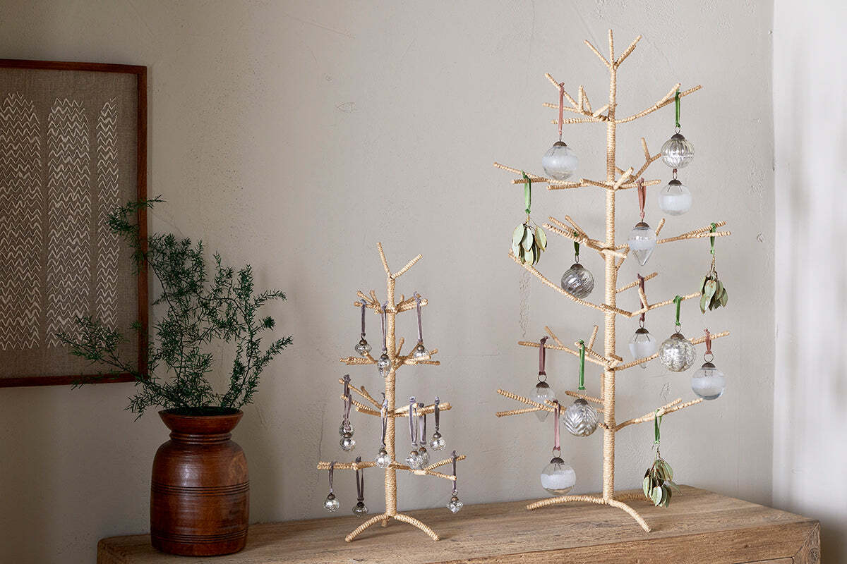 Two nkuku Mala Jute Trees in natural, with gold and woven jute branches, stand on a wooden surface. Each is adorned with glass ornaments and green accents, complemented by a brown vase of pine foliage and framed textile art for a festive look.