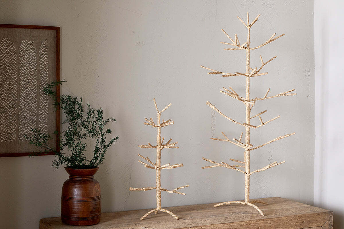 Two nkuku Mala Jute Trees, crafted from natural light jute, stand on a wooden surface beside a brown ceramic vase with green foliage. A light gray wall and framed art complete this beautifully minimalist festive decor.