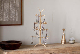 The nkuku Mala Jute Tree - Natural stands on a rustic shelf, paired with a metallic star-topped cone, a candle, wooden bowl, and framed textile art. The jute tree features gold tones and silver ornaments hung with gray ribbons.