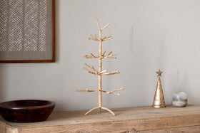 A minimalist gold wire tree sculpture stands on a rustic wooden surface beside nkuku’s Mala Jute Tree - Natural, a festive metallic cone with star topper, candle, and dark wooden bowl. The backdrop is a neutral wall with framed woven jute branches art.