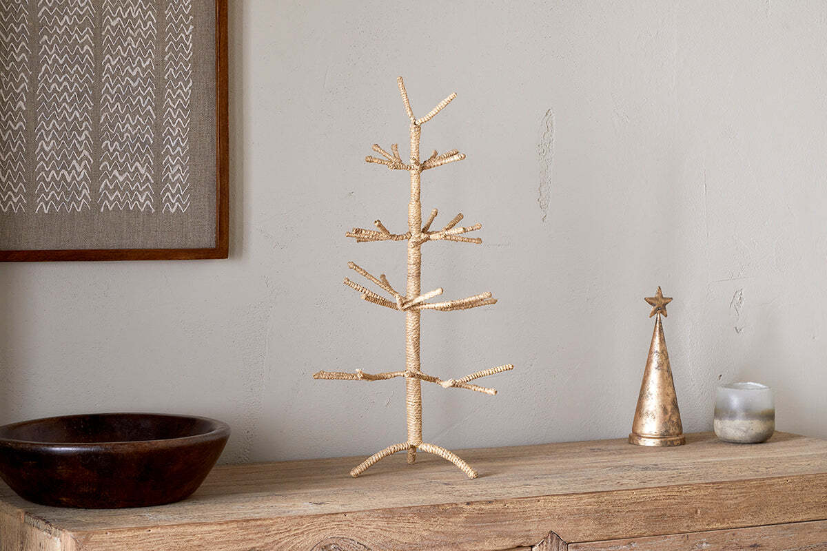 A minimalist gold wire tree sculpture stands on a rustic wooden surface beside nkuku’s Mala Jute Tree - Natural, a festive metallic cone with star topper, candle, and dark wooden bowl. The backdrop is a neutral wall with framed woven jute branches art.