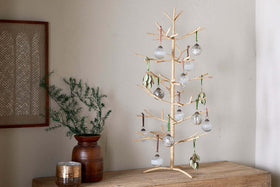 The Mala Jute Tree - Natural by nkuku, with minimalist woven jute branches, is displayed on a wooden surface, decorated with glass ornaments and green leaves. Beside it are a brown vase with greenery, a candle, and patterned artwork on a neutral wall.