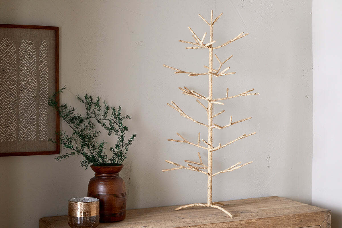 The Mala Jute Tree - Natural by nkuku stands on a wooden table beside a brown ceramic vase with green foliage, a decorative jar, and framed textile art, creating a festive display against the pale wall.