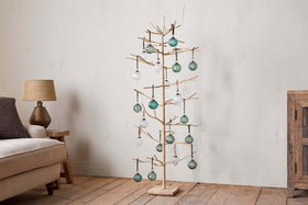 The nkuku Mala Base Jute Tree - Natural, with minimalist jute branches and gold accents, is adorned with green and clear glass ornaments, standing beside a beige sofa, rustic wooden cabinet, and table lamp against an off-white wall.