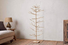 The nkuku Mala Base Jute Tree - Natural, with woven jute branches and a wooden base, stands on a wooden floor next to a beige sofa and small lamp table, set against a plain white wall in this cozy, rustic living room.