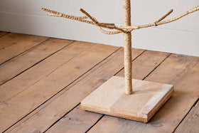 A close-up of the nkuku Mala Base Jute Tree - Natural shows its sisal-wrapped, branch-like arms and square wooden base, standing on a light wood floor beside a beige wall.
