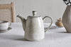 A nkuku Malia Teapot in Cream rests on a light tablecloth, accompanied by a matching teacup, a brown sugar jar, and a textured vase in the background. A woven chair is partially visible on the left.