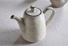 The nkuku Malia Teapot - Cream, featuring a cream-glazed stoneware body with subtle brown accents and a rounded lid, sits on a textured fabric surface. Part of another ceramic piece appears in the background.