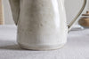 The nkuku Malia Teapot - Cream, made from cream-glazed stoneware with subtle speckles and textured vertical lines, rests on a light fabric surface. A cork-lidded jar appears softly blurred in the background.