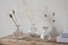 The Mayumi Recycled Glass Bud Vases - Clear (Set of 3) by nkuku are displayed with dried stems on a rustic wooden surface beside two stacked gray books, set against a textured light gray wall.
