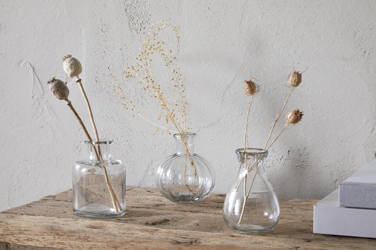 The Mayumi Recycled Glass Bud Vases - Clear (Set of 3) by nkuku are displayed with dried stems on a rustic wooden surface beside two stacked gray books, set against a textured light gray wall.