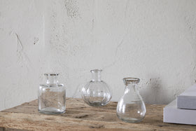 The nkuku Mayumi Recycled Glass Bud Vases – Clear (Set of 3) features three empty, uniquely shaped vases arranged on a rustic wooden surface against a textured off-white wall, with closed white books partially visible nearby.