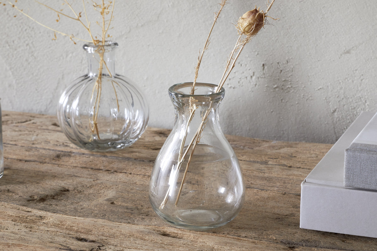The Mayumi Recycled Glass Bud Vases - Clear (Set of 3) by nkuku are displayed on a rustic wooden surface, with dried grasses in one vase and the others arranged nearby, creating an elegant set against a light textured wall.