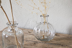The Mayumi Recycled Glass Bud Vases - Clear (Set of 3) by nkuku sit on a rustic wooden surface, each holding delicate dried stems, creating a minimalistic and natural look against a textured light gray wall.