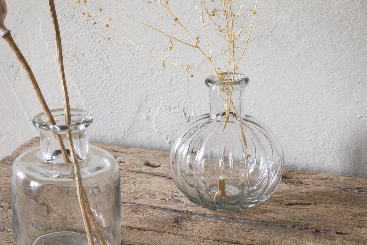The Mayumi Recycled Glass Bud Vases - Clear (Set of 3) by nkuku sit on a rustic wooden surface, each holding delicate dried stems, creating a minimalistic and natural look against a textured light gray wall.