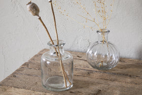 Three Mayumi Recycled Glass Bud Vases by nkuku sit on a rustic wooden surface, each holding dried botanicals. The clear glass vases are set against a textured, light-colored wall.