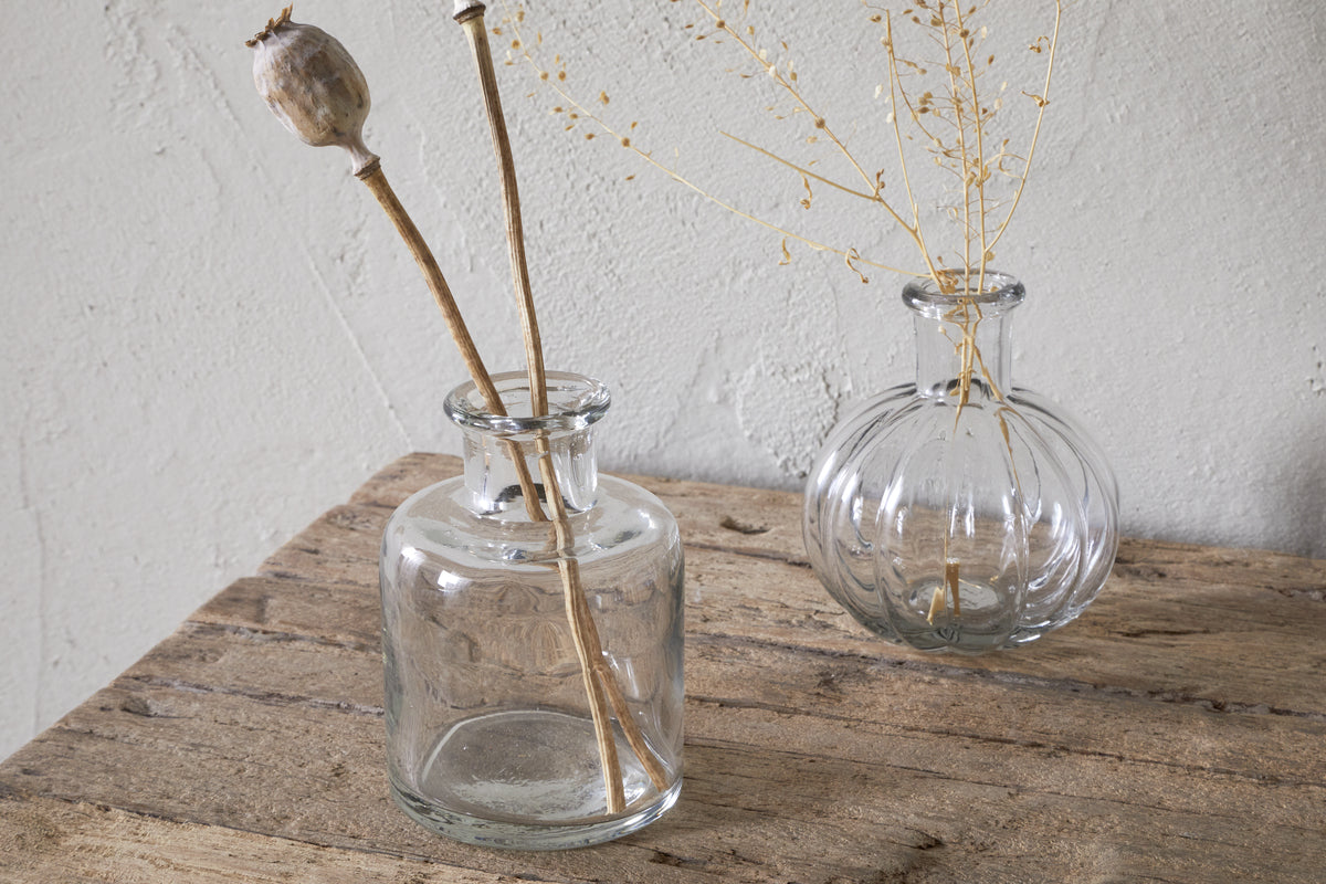 Three Mayumi Recycled Glass Bud Vases by nkuku sit on a rustic wooden surface, each holding dried botanicals. The clear glass vases are set against a textured, light-colored wall.