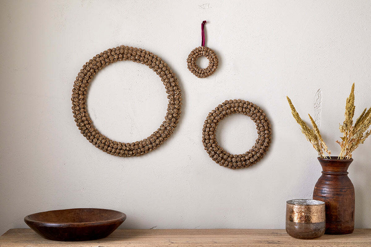 Three circular Madhi Wire Ball Wreaths in brass by nkuku hang on a light beige wall above a wooden surface, accompanied by a round wooden bowl, shiny candle holder, and brown vase with dried grasses for a rustic sculptural effect.