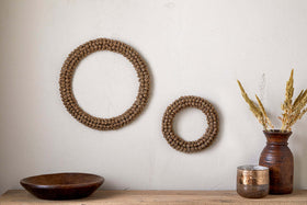 Two Madhi Wire Ball Wreaths by nkuku in brass create a striking sculptural decor display on a light beige wall. Below, a wooden bowl, brown vase with dried grasses, and metallic container complete the natural, rustic arrangement.