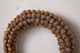 A close-up of the Madhi Wire Ball Wreath - Brass by nkuku, featuring tightly arranged brass wire balls in a layered design, elegantly displayed on a textured off-white wall.