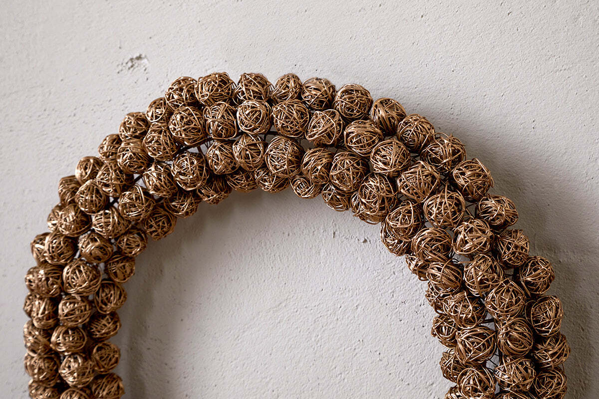 A close-up of the Madhi Wire Ball Wreath - Brass by nkuku, featuring tightly arranged brass wire balls in a layered design, elegantly displayed on a textured off-white wall.