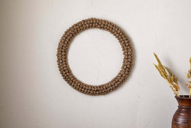 The Madhi Wire Ball Wreath in brass by nkuku hangs on an off-white wall. To the right, a ceramic vase with dried grasses adds a minimalist, rustic touch to this sculptural decor scene.