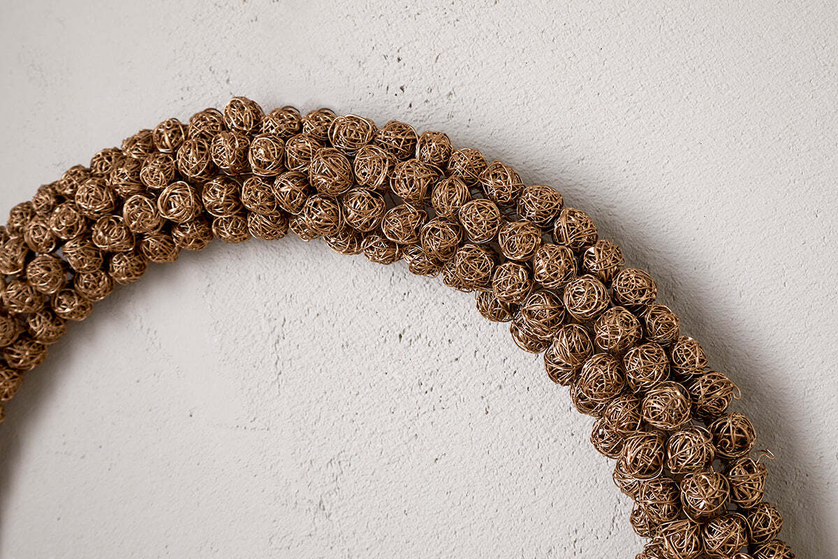 A close-up shows the Madhi Wire Ball Wreath in brass by nkuku, featuring tightly woven wire balls. Part of the arc is visible against a textured light wall, creating a striking sculptural display.