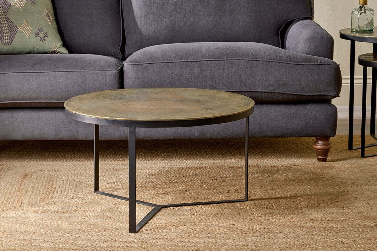 The nkuku Maba Coffee Table, featuring a bronze top and slim black legs, stands on a beige woven rug before a dark gray sofa with wooden legs and a green cushion. A small round side table is partly visible on the right.