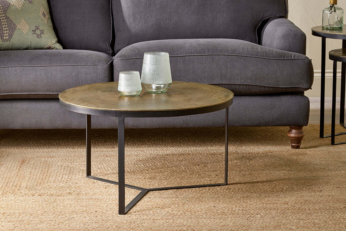 The Maba Coffee Table by nkuku, featuring a metallic top and black geometric legs, sits on a woven rug before a gray sofa. Two glass vases and a patterned pillow complete this charming and inviting living room setup.