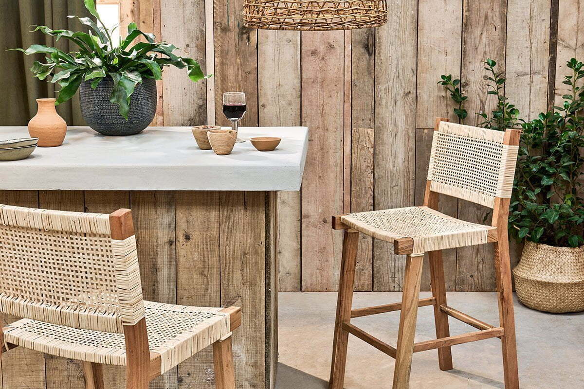 A rustic kitchen features the Madrisana Acacia & Rattan Bar Stool by nkuku, a white countertop with pottery, a glass of red wine, and a potted plant. Wooden plank walls and green plants in woven baskets enhance the cozy atmosphere.