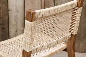 A close-up of the Madrisana Acacia & Rattan Bar Stool by nkuku, featuring a woven cane seat and backrest on a light brown acacia frame. Wrapped cane details accent the joints, set against rustic, vertically arranged wooden planks in the background.