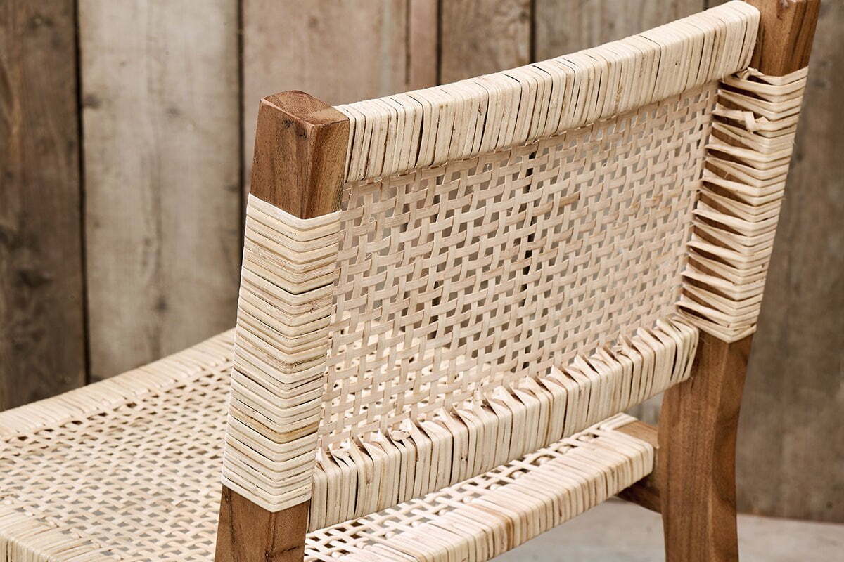 A close-up of the Madrisana Acacia & Rattan Bar Stool by nkuku, featuring a woven cane seat and backrest on a light brown acacia frame. Wrapped cane details accent the joints, set against rustic, vertically arranged wooden planks in the background.