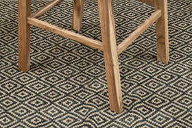 Close-up of the Madrisana Acacia & Rattan Bar Stool by nkuku, showing its natural-finish legs on a black and beige diamond-patterned woven rug—highlighting the contrast between the stool’s sturdy frame and the geometric design below.