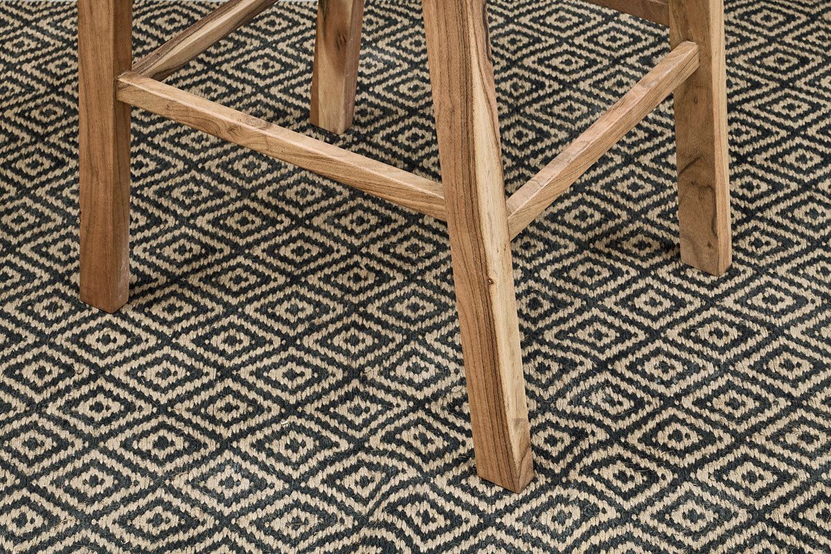 Close-up of the Madrisana Acacia & Rattan Bar Stool by nkuku, showing its natural-finish legs on a black and beige diamond-patterned woven rug—highlighting the contrast between the stool’s sturdy frame and the geometric design below.