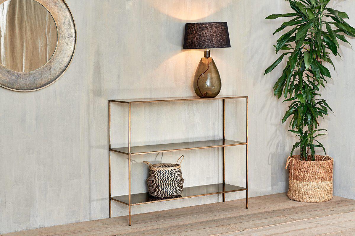 The nkuku Mahi Console Table, with sleek brass shelves, stands by a light gray wall. It displays a brown glass lamp with black shade and a woven basket, beside a tall potted plant and a round wall mirror.
